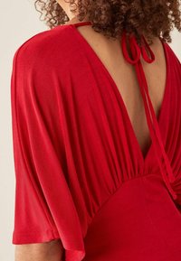 Robe rouge avec manches courtes et fluides, dos largement ouvert. Taille froncée et fermeture par lien au niveau du cou. Tissu doux et texturé.