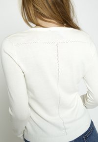 Donna con capelli castano chiaro che indossa un maglione bianco a maniche lunghe con piccole linee perforate verticali e orizzontali sulla schiena e jeans blu.