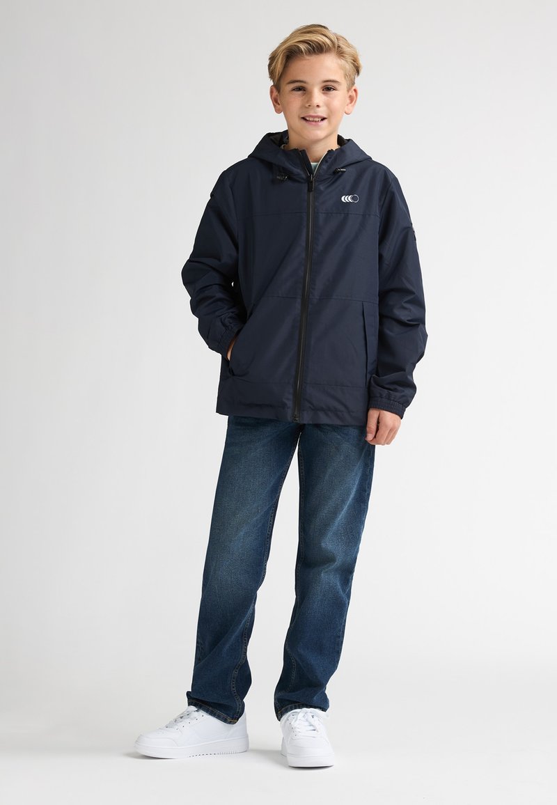 Veste coupe-vent bleu marine avec une fermeture éclair à l'avant, des poignets élastiques et une capuche à cordon de serrage. Portée avec un jean bleu et des baskets blanches, avec des poches latérales.