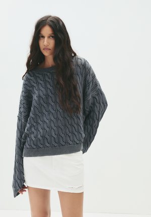 Anthrazitgrauer Strickpullover mit Kabelmuster, lockere Passform, weite Ärmel und geripptem Saum, kombiniert mit einem weißen Denim-Minirock.