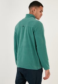 Giacca in pile verde con colletto alto, caratterizzata da una texture liscia e un piccolo logo nero sulla parte posteriore, abbinata a pantaloni scuri.