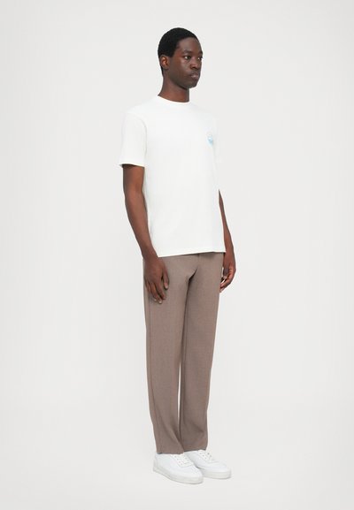 Hvid kortærmet t-shirt med et blåt logo, parret med lysebrune skræddersyede bukser og hvide sneakers, stående mod en ensfarvet baggrund.