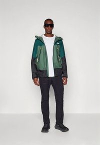 Green and black outdoor jacket with zipper and hood, paired with black pants and dark shoes. Model wears sunglasses and a white shirt.
