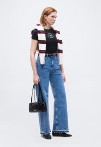 Femme aux cheveux courts debout portant un t-shirt noir, une écharpe rayée sur les épaules, un jean taille haute, des chaussures noires, et tenant un sac à main noir.