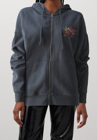 Felpa zip-up grigio scuro realizzata in tessuto morbido, con tasca frontale e un design ricamato di un cuore e una rosa sul lato sinistro del petto.
