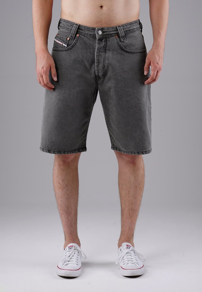 Male wearing grey denim shorts and white low-top sneakers standing against a plain grey background.