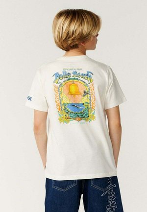 Blondhaarige Person trägt ein weißes T-Shirt mit farbenfrohem Hello Beach-Motiv und blaue Jeans mit weißem Text auf den Gesäßtaschen.