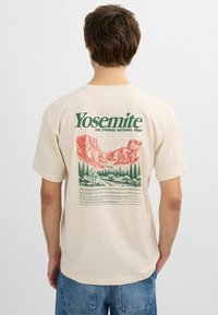 Persona con camiseta beige con gr�áfico y texto en rojo y verde del Parque Nacional Yosemite en la parte trasera, combinado con jeans de mezclilla azul.