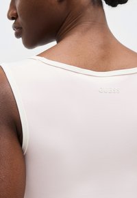 Κοντινό πλάνο στο πάνω μέρος της πλάτης και στον ώμο ενός ατόμου που φοράει αμάνικη κορυφή σε ανοιχτό μπεζ χρώμα με το λογότυπο «GUESS» ανάγλυφο κοντά στο λαιμό.