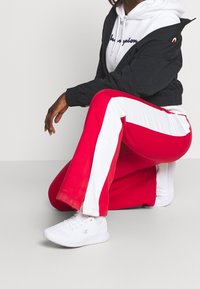 Persona in ginocchio che indossa pantaloni da jogging rossi e bianchi, sneakers bianche, felpa bianca e una giacca nera corta su uno sfondo neutro.