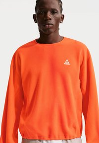 Άνδρας με κοντά πλεξούδες φοράει φωτεινό πορτοκαλί φλις με μακριά μανίκια και λευκό τριγωνικό λογότυπο ACG στο στήθος, στέκεται απέναντι σε λευκό φόντο.