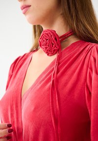 Choker en tissu rouge avec un design de rose texturé et superposé. Le choker se fixe autour du cou avec de fines lanières assorties, complétant une robe rouge.
