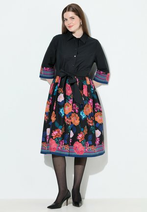 Schwarzes Kleid mit tailliertem Oberteil, floralen Mustern in lebhaften Farben auf dem Rock und gestreiften Akzenten an den Ärmeln. Mit einer gebundenen Taillenpartie, knielang.
