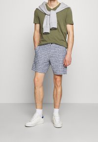 Camiseta verde de corte en V, pantalones cortos grises con un patrón geométrico, suéter gris claro colocado sobre los hombros, zapatillas blancas y calcetines de longitud media.