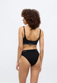 Μαύρο ασύρματο σουτιέν με ρυθμιζόμενους ιμάντες και λωρίδα με λογότυπο· συνδυασμένο με μαύρο σλιπ ψηλού κόψιμου, και τα δύο από λείο υλικό.