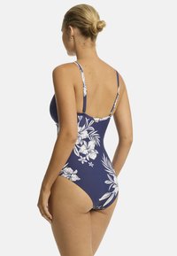Model gezien van de achterkant, gekleed in een marineblauwe badpak met witte bloemmotieven en dunne bandjes, met een gladde silhouet.