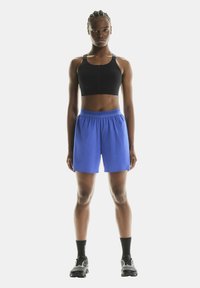 Schwarzer Sport-BH mit Reißverschluss vorne, kombiniert mit leuchtend blauen Sportshorts mit elastischem Bund, getragen mit schwarzen Socken und Sneakers.