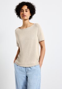 Beige kortärmad topp med vid halsringning, tillverkad av mjukt, texturerat tyg. Parats med ljusblå, löst sittande byxor.