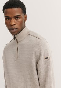 Beige Kapuzenpullover mit Reißverschluss, strukturiertem Muster, hohem Kragen und subtiler Markenkennzeichnung am Ärmel. Slim-Fit mit gerippten Details.