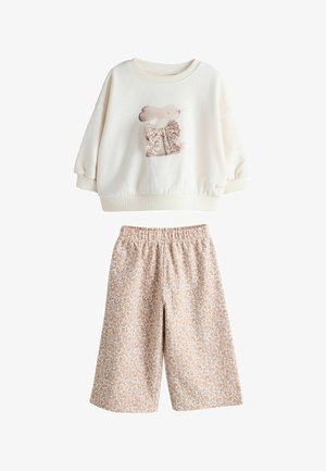 Šviesiai kreminės spalvos bliuzonas su triušio grafika ir gėlėtu lankeliu, derinamas su smėlio spalvos šortais su gėlių raštu. Abu drabužiai laisvo kirpimo.