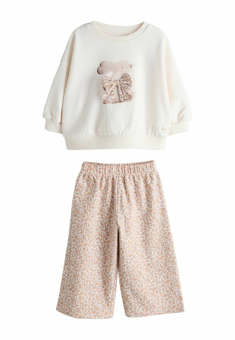 Hellcremiger Pullover mit Hasenmotiv und Blumenschleife, kombiniert mit beigen Shorts mit Blumenmuster. Beide Kleidungsstücke haben eine lockere Passform.