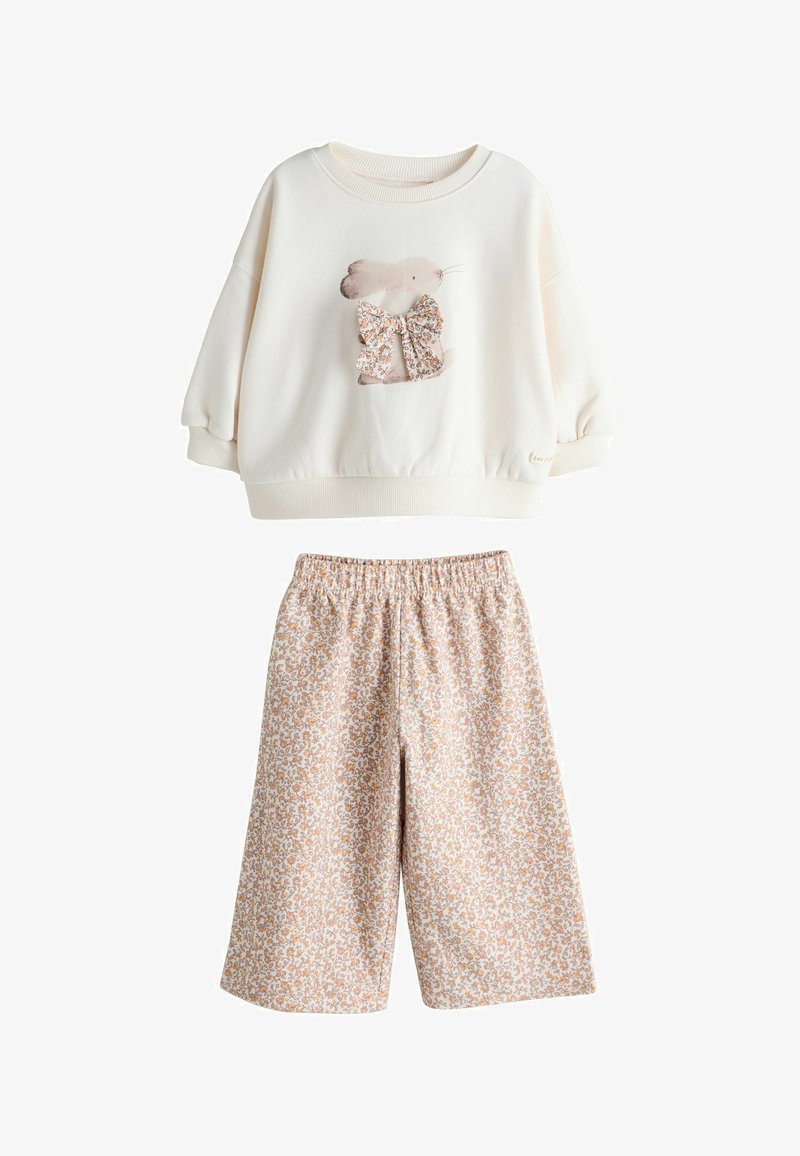 Hellcremiger Pullover mit Hasenmotiv und Blumenschleife, kombiniert mit beigen Shorts mit Blumenmuster. Beide Kleidungsstücke haben eine lockere Passform.