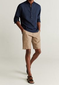 Camicia a maniche lunghe in lino blu navy con colletto, abbinata a pantaloni corti in lino beige chiaro e sandali in pelle marrone.