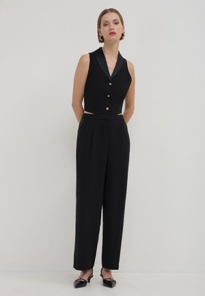 Femme portant un gilet sans manches noir avec des boutons et un pantalon noir à larges jambes taille haute, se tenant les mains derrière le dos.