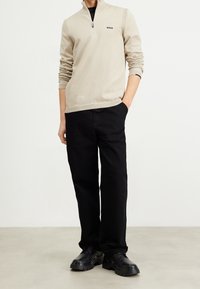 Pull-over beige à demi-fermeture éclair avec des manches texturées et un logo noir, associé à un pantalon ample noir et des chaussures noires épaisses.
