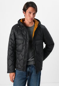 Casaco preto acolchoado com capuz amarelo em contraste, fecho de zipper e dois bolsos no peito. O tecido parece suave e leve.