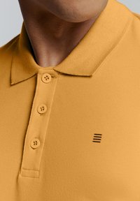 Mosterdgele polo shirt van een textuurstof, met drie knopen en een klein zwart gestreept logo op de linkerborst.