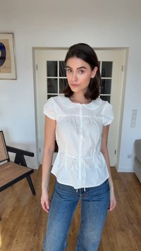 Femeie în picioare în interior, purtând o bluză albă cu mâneci scurte și nasturi și blugi albaștri, cu podea din lemn și un scaun în fundal.