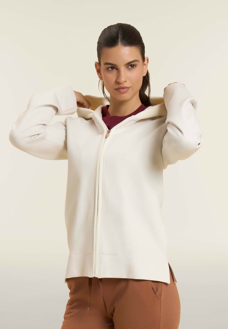 Felpa con zip di colore crema, con cappuccio regolabile, polsini a costine e fessure laterali. Indossata sopra una maglietta bordeaux, con una texture liscia.