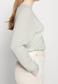 Kvinna i en ljusgrå, texturerad tröja med långa ärmar, instoppad i högmidjade beige byxor med ett matchande bälte, hand i fickan.