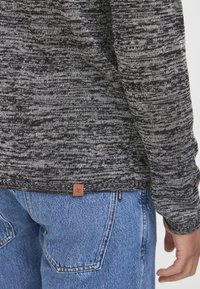 Zwart en grijs gemêleerd brei-trui, met een bruine leren merklabel aan de zoom, gecombineerd met blauwe denim jeans met opgestikte zakken.