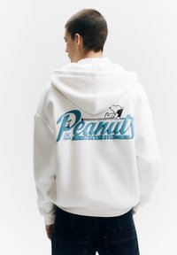 Witte hoodie met een grote blauwe print "Peanuts Hockey Club" op de achterkant, met een afbeelding van Snoopy. De stof lijkt zacht en gestructureerd.