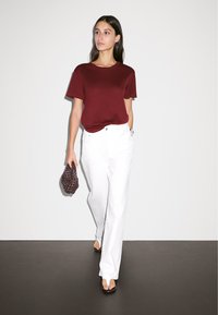 Burgundy cotton t-shirt, white straight-leg jeans, and brown flip-flops. Model holds a textured, dark woven bag. Minimal background.
