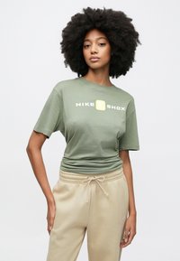 Πράσινο ελιάς βαμβακερό t-shirt με το λογότυπο "NIKE SHOX" σε λευκό και κίτρινο, συνδυασμένο με μπεζ φόρμα με κορδόνι στη μέση και πλαϊνή τσέπη.