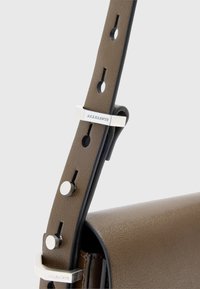 Ruskea nahkainen laukkuhihna, jossa on säädettävät rei'ät, hopeiset metalliosat ja merkkityöstö metallisoljessa. Sileä pinta.