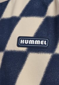 Beige tyg med ett marinanavyrutat mönster, med en rektangulär svart etikett med vit text "HUMMEL".