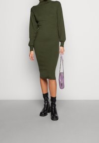 Robe en maille vert olive avec un col montant et de longues manches bouffantes. Associée à des bottines noires et un petit sac à main lavande.