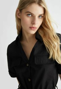 Camicia nera con bottoni realizzata in tessuto leggero, dotata di colletto, maniche corte e due tasche frontali. Texture liscia con una vestibilità rilassata.