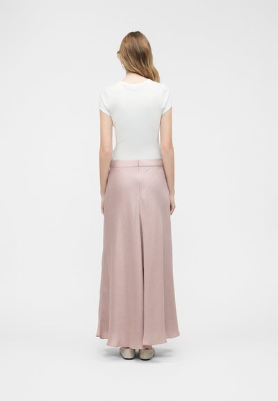 Γυναίκα με μακριά μαλλιά που φοράει λευκό κοντομάνικο τοπ και αέρινη μακριά φούστα σε απαλό ροζ χρώμα, στέκεται με την πλάτη προς το μέρος μας σε απλό φόντο.