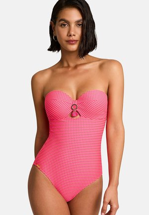 Femme portant un maillot de bain une pièce rose texturé sans bretelles avec un anneau métallique central et un détail pompon, debout devant un fond uni.
