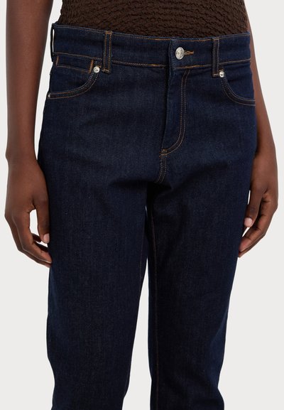 Σκούρο μπλε τζιν από τζιν ύφασμα, με κλασικό σχέδιο πέντε τσεπών, φερμουάρ και αντίθετες πορτοκαλί ραφές. Λείο υλικό.