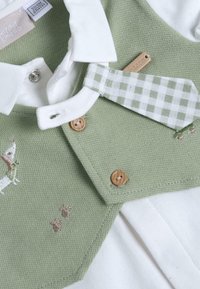 Gilet verde a maglia con colletto bianco, bottoni di legno e cravatta a scacchi. Presenta disegni ricamati di animali e materiale in cotone morbido.