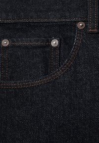 Mørkeblåt denimstof med close-up af en lille forlommel, sikret med tre metalnitter og brune sømdetaljer.