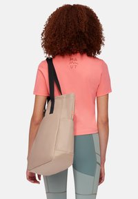 Sac fourre-tout en tissu beige avec des poignées noires, comportant une poche latérale. Porté avec un t-shirt corail et des leggings verts. Pas de motifs visibles.