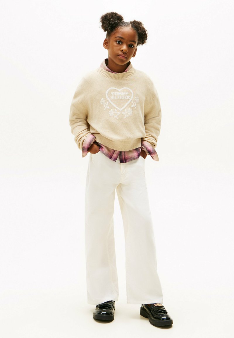 Beiger Pullover mit weißem Herz-Logo, übereinander getragen mit einem rosa karierten Hemd. Weiße weit geschnittene Hose und schwarze Schuhe mit strukturiertem Design.