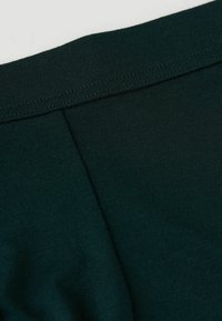 Tmavě zelená látka zblízka s hladkým povrchem, s žebrovaným pasem a jemným švem, ideální pro volnočasové nošení.